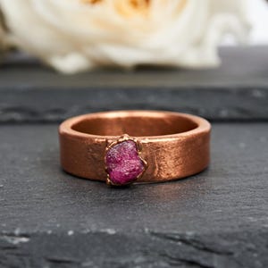 May include: A copper ring featuring a textured, metallic band with a prominent, raw, deep pink gemstone set at the center. The metal surface displays a warm, reddish-brown tone with visible handcrafted markings and a slightly uneven, organic finish.