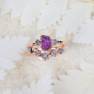 Puede incluir: Un conjunto de dos anillos de oro rosa. El anillo superior presenta una piedra preciosa púrpura en bruto central flanqueada por dos pequeñas piedras grises. El anillo inferior tiene forma de V y está engastado con cinco piedras grises.