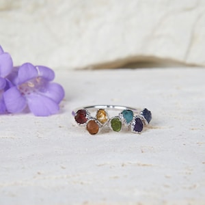 May include: A silver ring featuring a row of seven small, raw, multi-coloured gemstones including red, orange, yellow, green, blue, and purple stones set along the top of the band. The ring is positioned next to a cluster of purple flowers.