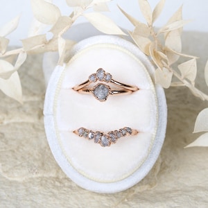 May include: Two rose gold rings featuring raw, grey, uncut diamonds. The top ring consists of a central diamond flanked by two smaller stones, while the bottom ring is a curved band set with a series of seven small, irregular grey diamonds.