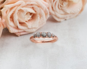 May include: A rose gold ring featuring five small, rough, grey, uncut diamonds set in a row along the top of the band. The metal band has a warm, metallic copper-toned finish, and the stones have a textured, natural, and irregular appearance.