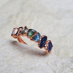 May include: A ring featuring a row of six raw, uncut gemstones in a gradient of colours ranging from light green to deep blue, set in a textured rose gold band. The stones are irregular in shape and size, creating a natural, organic appearance.