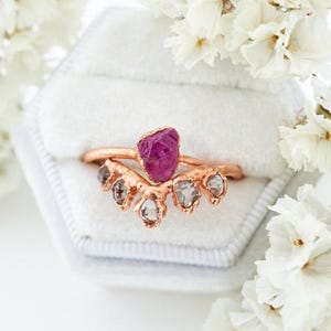 May include: A rose gold ring featuring a central raw, deep pink gemstone flanked by four smaller, translucent, irregular-shaped stones arranged in a chevron pattern. The metal band has a textured, organic finish and rests in a white velvet box.