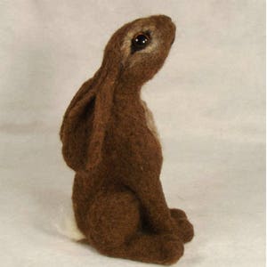 Puede incluir: Una escultura de conejo de fieltro de aguja marrón y blanco con ojos marrones. El conejo está sentado erguido con las orejas levantadas.