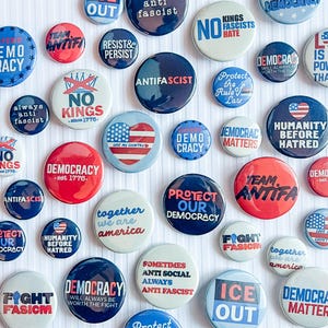 May include: A collection of round buttons with various political slogans and phrases. The buttons feature text such as "Democracy," "Anti-fascist," and "Love is More Powerful Than Hate." Colors include red, white, and blue.