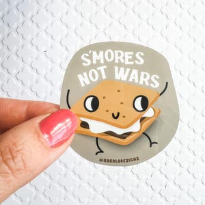 May include: A round sticker with a cartoon s'more with googly eyes and a smiling mouth. The s'more is holding a sign that says "S'mores Not Wars" in white text on a gray background.