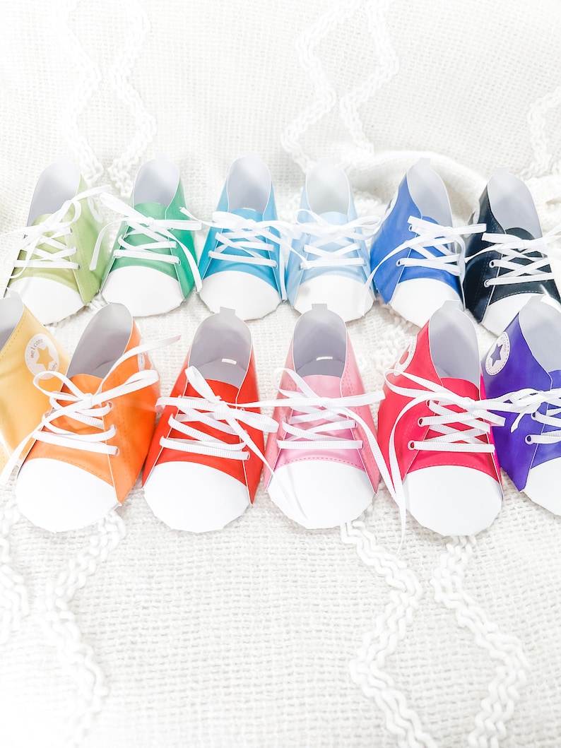 May include: A set of ten paper baby shoes in various colors, including green, blue, yellow, orange, red, pink, purple, and black. The shoes are decorated with white laces and a white sole.