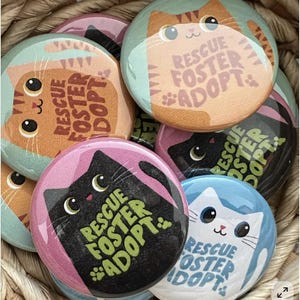 May include: A collection of circular pin-back buttons featuring stylized cat illustrations in various colors including orange, black, and white. Each button displays the text "RESCUE FOSTER ADOPT" accompanied by small paw print graphics, showcasing a variety of feline designs.