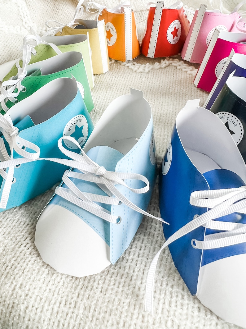 May include: A set of colorful paper baby shoes with white laces. The shoes are decorated with a white star and the text "Welcome Baby".
