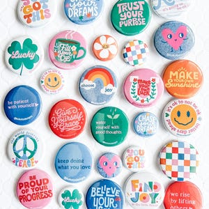 May include: A collection of round pin-back buttons featuring various colorful designs, including inspirational text like 'You Got This', 'Chase Your Dreams', 'Trust Your Purpose', 'Lucky', 'Make Your Own Sunshine', 'Be Patient With Yourself', and 'Find Joy'.