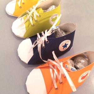 May include: Three miniature Converse-style shoes made of paper, each in a different color: yellow, green, and blue. Each shoe has a white toe and laces, and a small circular sticker with the text "Welcome Baby" on the side. The shoes are filled with chocolate kisses.