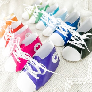 May include: A row of colorful paper shoe-shaped party favors with white laces and a white star on the side. The shoes are arranged in a rainbow pattern from gold to black.