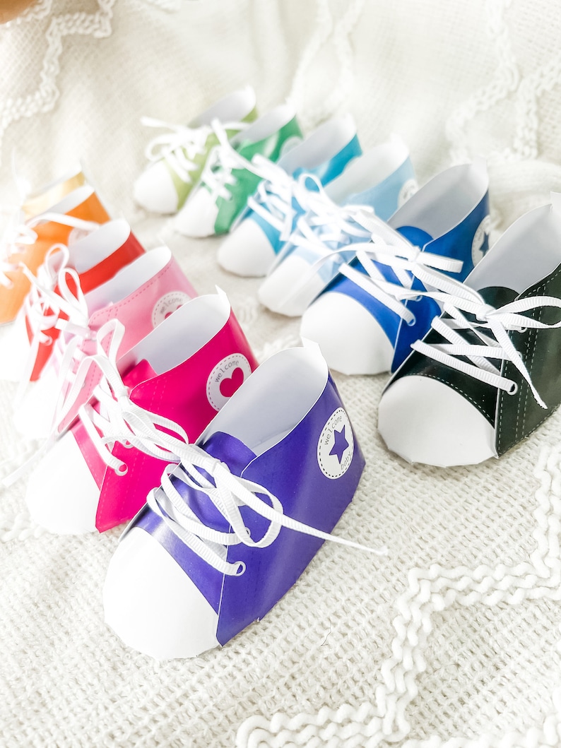 May include: A row of colorful paper shoe-shaped party favors with white laces and a white star on the side. The shoes are arranged in a rainbow pattern from gold to black.