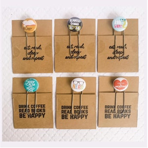 May include: Six different book marks with various designs. Each bookmark is a brown card with a gold binder clip and a round button with a design. The buttons feature designs such as "Readers Gonna Read", "Keep Calm and Read On", "Nerd", "Rather be Reading", "Worm", and "One More Chapter".