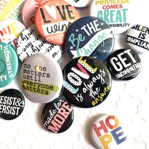 May include: A collection of colorful button pins with various messages, including "Love is always the answer", "Be the Change", "No one matters until everyone matters", "Get uncomfortable", "Hope", "Made for More", and "Love wins every time".