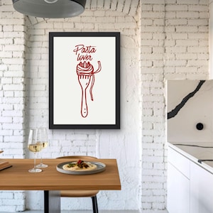 May include: A framed print featuring a red illustration of a fork twirling spaghetti, with the words "Pasta lover" in cursive. The artwork is set against a cream background, framed in black, and hangs on a white brick wall.
