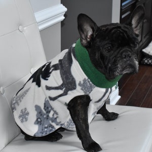 May include: A black French Bulldog wearing a white fleece dog coat with a gray reindeer pattern and a green collar. The dog is sitting on a white chair, looking towards the right.