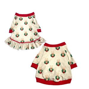 May include: Two beige dog sweaters with a red trim and a reindeer pattern. The sweater on the left has a ruffled hem.