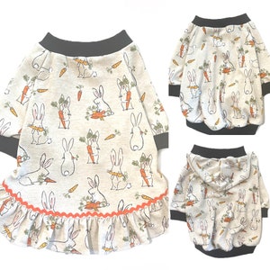 May include: A beige dog sweater with a ruffle trim and a pattern of bunnies holding carrots. The sweater is available in two styles: a hooded sweater and a sweater with a crew neck.