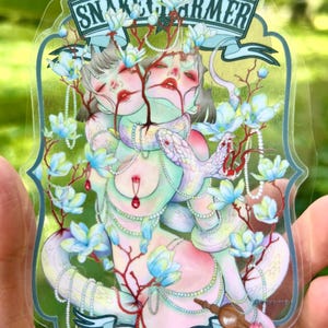 Könnte beinhalten: Ein durchsichtiger Aufkleber mit einer zweiköpfigen Figur, die von einer weißen Schlange umschlungen ist, verziert mit Perlenketten und blauen Magnolienblüten. Das Design enthält rote Zweigakzente und Banner mit dem Text SNAKE CHARMER und FREAK SHOW.