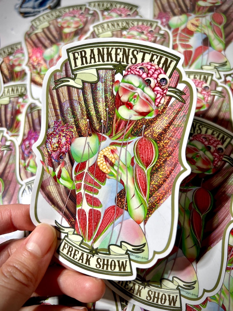 K&ouml;nnte beinhalten: Ein gestanzter Aufkleber mit einer stilisierten anatomischen Figur mit freiliegenden Muskeln und einem Gehirn, die ein Herz h&auml;lt. Das Design enth&auml;lt den Text FRANKENSTEIN oben und FREAK SHOW unten, mit glitzerndem Finish und feinen Linien.