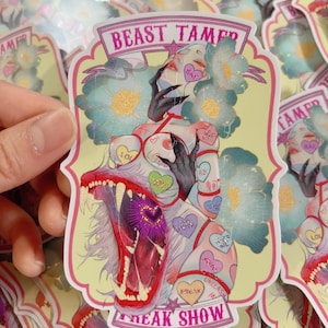 Könnte beinhalten: Ein Aufkleber mit gelbem Hintergrund. Es zeigt eine stilisierte Illustration einer Frau mit schwarzen Krallen und einem Wolfsmaul. Die Texte "BEAST TAMER" und "FREAK SHOW" sind abgebildet. Die Frau hat blaue Blumenakzente und herzförmige Tattoos.