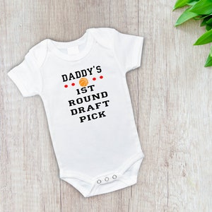 May include: White baby bodysuit with the text "DADDY'S 1ST ROUND DRAFT PICK" in black and red. A basketball graphic is centered above the text. The bodysuit has short sleeves and poppers at the bottom. The background is a light wood surface.