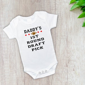 May include: White baby bodysuit with the text "Daddy's 1st Round Draft Pick" in black and red. The bodysuit features a football graphic and is designed for a newborn. The garment has popper closures at the bottom.