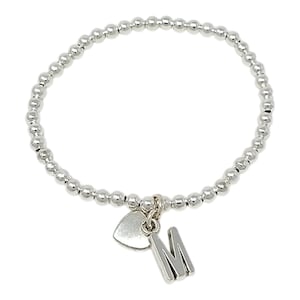 May include: A silver-toned stretch bracelet composed of small, uniform spherical beads. The bracelet features two dangling charms: a smooth, solid heart-shaped charm and a capital letter M charm, both finished in a matching polished silver tone.