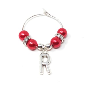 May include: A silver wine charm with a red bead design. The charm features a silver letter "R" pendant and is accented with clear rhinestones. The beads are a glossy red colour.