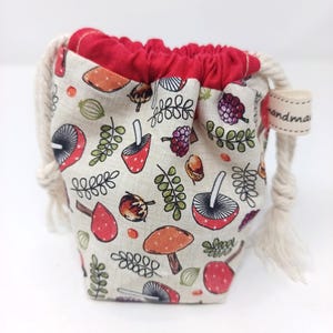 May include: A small fabric drawstring pouch featuring a beige base with a repeating pattern of red and orange mushrooms, green leaves, and purple berries. The pouch has a bright red interior lining and thick cream-coloured rope ties with a handmade tag.