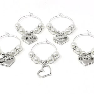 May include: Set of four silver wine glass charms with white pearl beads. Each charm features a heart-shaped tag with the words "Bride", "Groom", "Bridesmaid", and "Flower Girl".