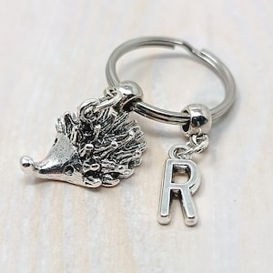 May include: A silver-toned metal keyring featuring a three-dimensional hedgehog charm and a separate letter R charm attached to a circular split ring. The hedgehog charm has detailed quills and a smooth snout, while the letter R is a simple block font.