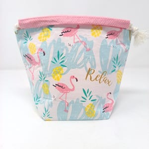 May include: A fabric pouch featuring a light pink, light blue, and yellow pattern of flamingos and pineapples. The word Relax is printed in gold script on the front. The top edge has a pink polka dot trim and a white drawstring closure.