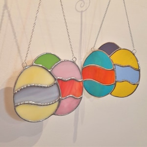 May include: Two stained glass ornaments hanging from silver chains. The ornament on the left is made of four pieces of glass in shades of yellow, purple, pink, and green. The ornament on the right is made of three pieces of glass in shades of blue, orange, and yellow.