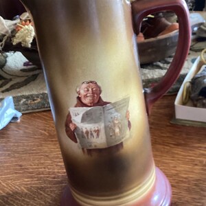 May include: A brown and white ceramic pitcher with a red handle. The pitcher features a painted image of a man wearing glasses and a robe, reading a newspaper.