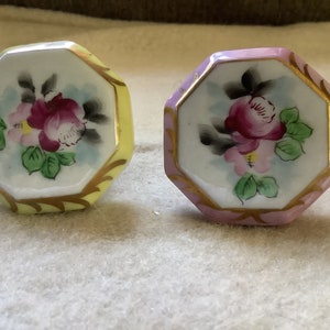 May include: Two octagonal porcelain cabinet knobs. One is yellow, the other is pink. Each features a hand-painted floral design with a central burgundy rose, pink accents, and green leaves. Gold trim outlines the edges.