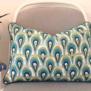 May include: Decorative rectangular pillow with a teal, blue, and green peacock feather pattern. The pillow has a white background with vertical stripes and a teal border. The pillow is placed on a gray chair.