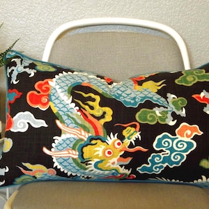 May include: Rectangular decorative pillow with a black background and a colorful dragon design. The dragon is primarily blue, yellow, and red, with accents of green and white. The pillow has a blue border and is placed on a chair.