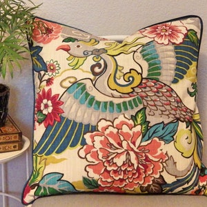 May include: A decorative pillow with a white background and a colorful design featuring a stylized bird with blue, green, yellow, and gray feathers. The bird is surrounded by pink and red flowers with green leaves.