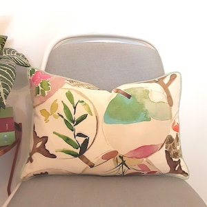 May include: Rectangular decorative pillow with a cream background and a watercolor floral design. The pillow features green, pink, and brown botanical elements, with a light blue border. The pillow is placed on a gray chair.