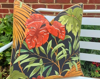 May include: A black pillow with a tropical print featuring large red flowers, green leaves, and gold accents. The pillow has a gold trim around the edges.