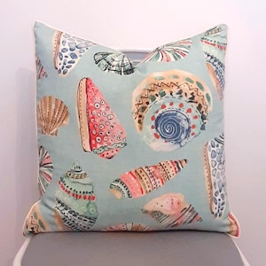 May include: Square decorative pillow with a light blue background and a pattern of colorful seashells. The seashells are in shades of pink, red, green, and brown, with intricate details. The pillow has a white border.