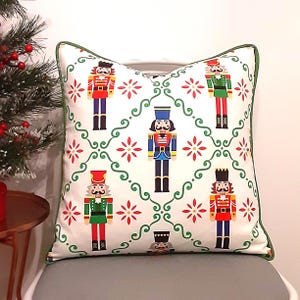May include: A square throw pillow with a white background featuring a repeating pattern of colorful nutcracker figures in red, blue, and green uniforms, interspersed with red floral motifs and green swirling lines, finished with green piping.