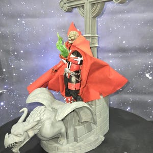 Könnte beinhalten: Detaillierte Actionfigur eines Charakters mit rotem Umhang, schwarzer und roter Rüstung und einem schädelartigen Gesicht, der grüne Flammen hält. Die Figur steht auf einer Steinstruktur mit einem Wasserspeier und einem Kreuz.