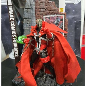 Puede incluir: Figura de acción de Spawn con capa roja, traje blanco y negro y cadenas. La figura está posada sobre una base detallada con elementos de ladrillo y piedra. El embalaje incluye el texto "IMAGE COMICS FOUNDERS".