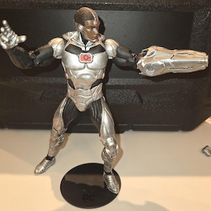 Puede incluir: Una figura de acción de Cyborg, un superhéroe de DC Comics, en color plateado y negro. La figura está en una postura de acción dinámica con un brazo extendido hacia adelante. La figura tiene un ojo rojo y un símbolo rojo en el pecho. La figura está de pie sobre una base negra con el logotipo de DC Comics.