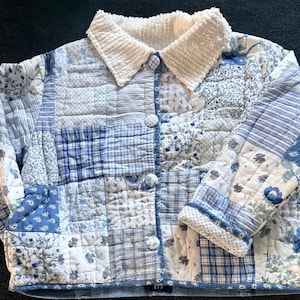 May include: A quilted jacket in shades of blue and white, featuring a patchwork design. The jacket has a corduroy collar and button closures. The sleeves and body are composed of various floral and geometric patterns. The jacket is a cozy, handmade garment.