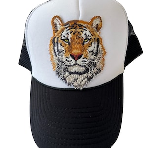 May include: A black and white trucker hat featuring an embroidered tiger face. The tiger has orange and brown fur with green eyes and a pink nose. The hat has a black brim and mesh backing.