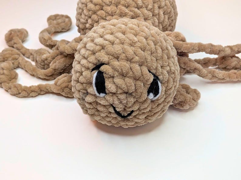 A close up of the face of a light brown fidget toy spider. They eyes have been embroidered with black yarn and white highlights added. There is also a small smile and some eyebrows that give the spider a plaintive look.